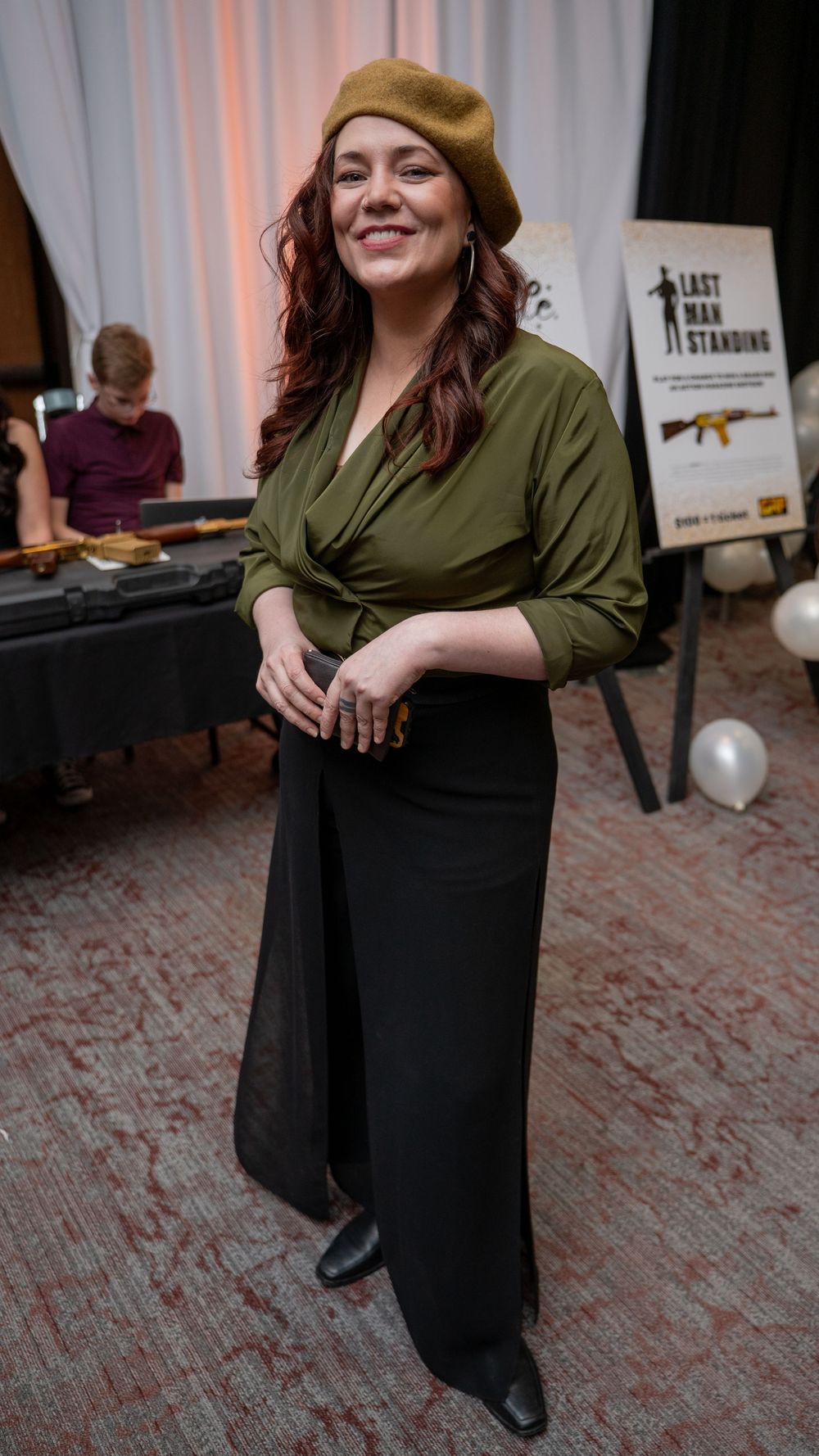 Woman in olive green blouse, black skirt, and gold beret smiles at an indoor event near a Last Man Standing poster.