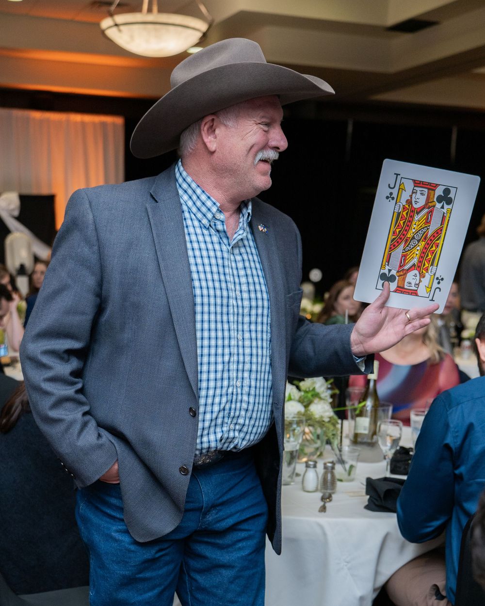 Man in cowboy hat and blazer holds a King of Clubs playing card at an indoor event.