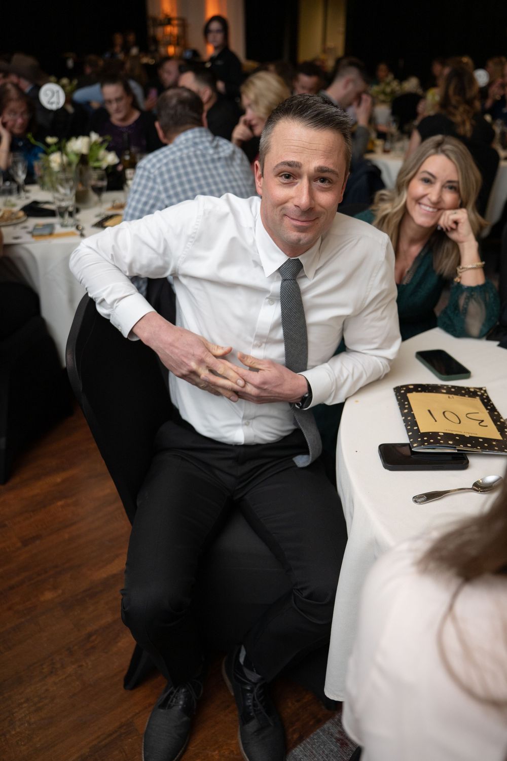 A man in a white shirt and gray tie smiles at a formal banquet dinner event with guests seated at white tablecloth tables.