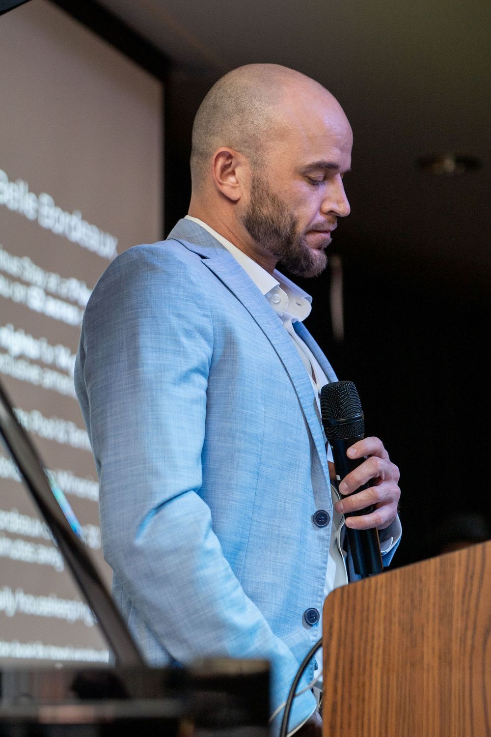 Bald man with beard in light blue blazer speaking at podium holding microphone at professional conference event.