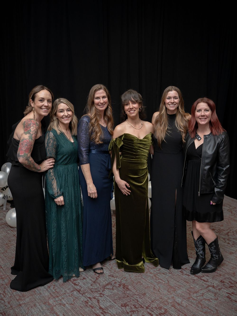 Seven women in elegant formal gowns and cocktail attire pose together smiling at a glamorous event.