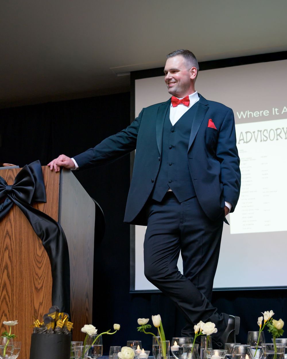 Man in tuxedo with red bow tie speaking at formal event with presentation screen behind him.