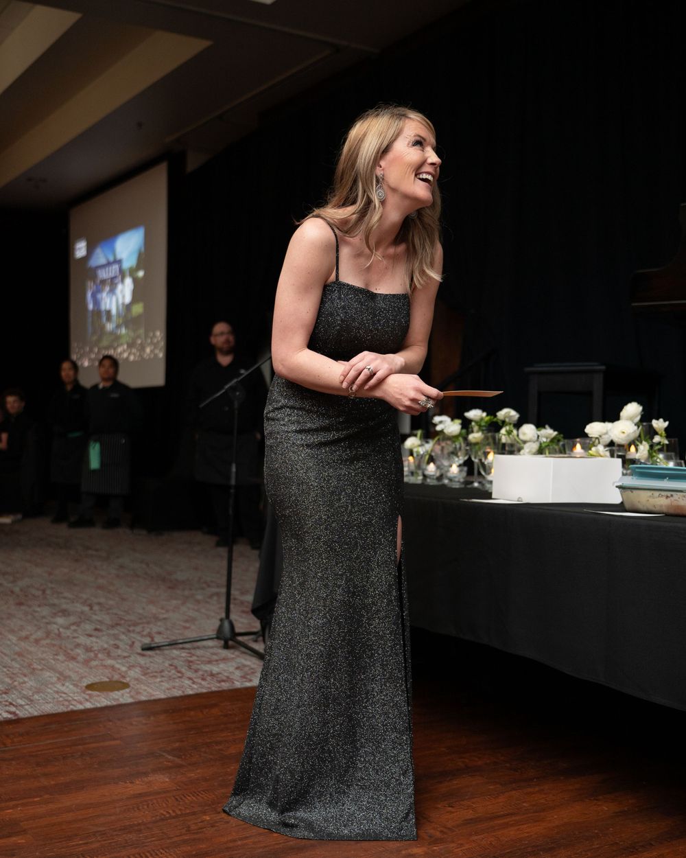 Woman in elegant black sparkly gown smiling at formal gala event with floral arrangements and presentation screen.
