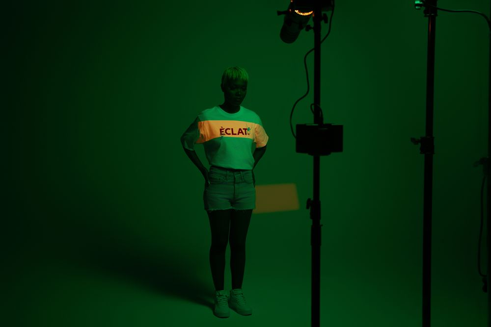 A person wearing a green t-shirt and jeans stands against a dark green background.