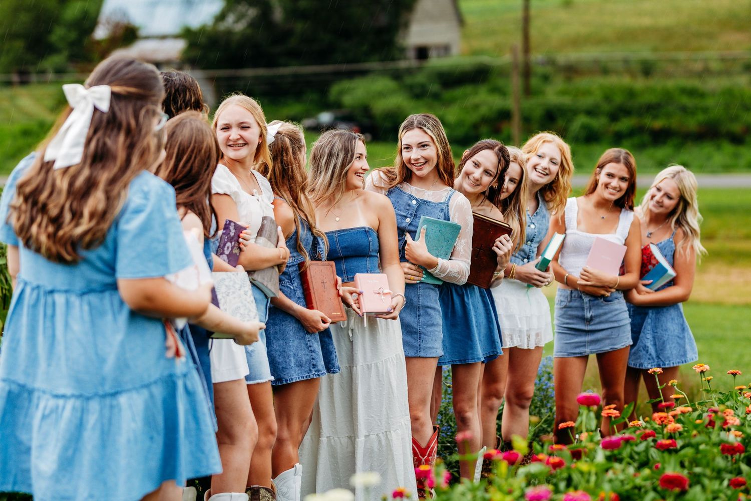 Group of smiling young women in blue dresses holding books in a colorful flower garden.