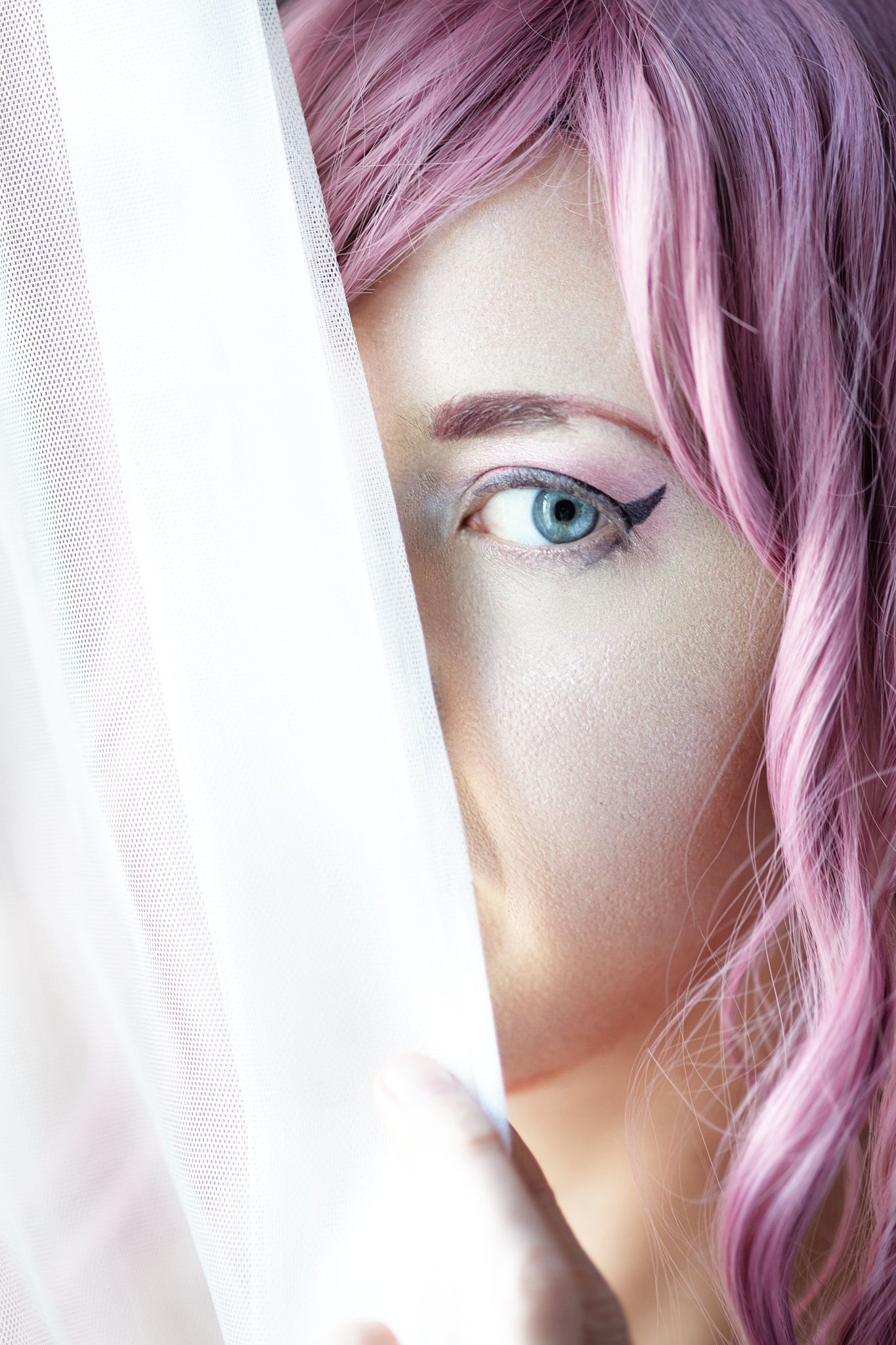 Close up portrait with pink hair and white curtains showing bright blue eyes and soft lighting.