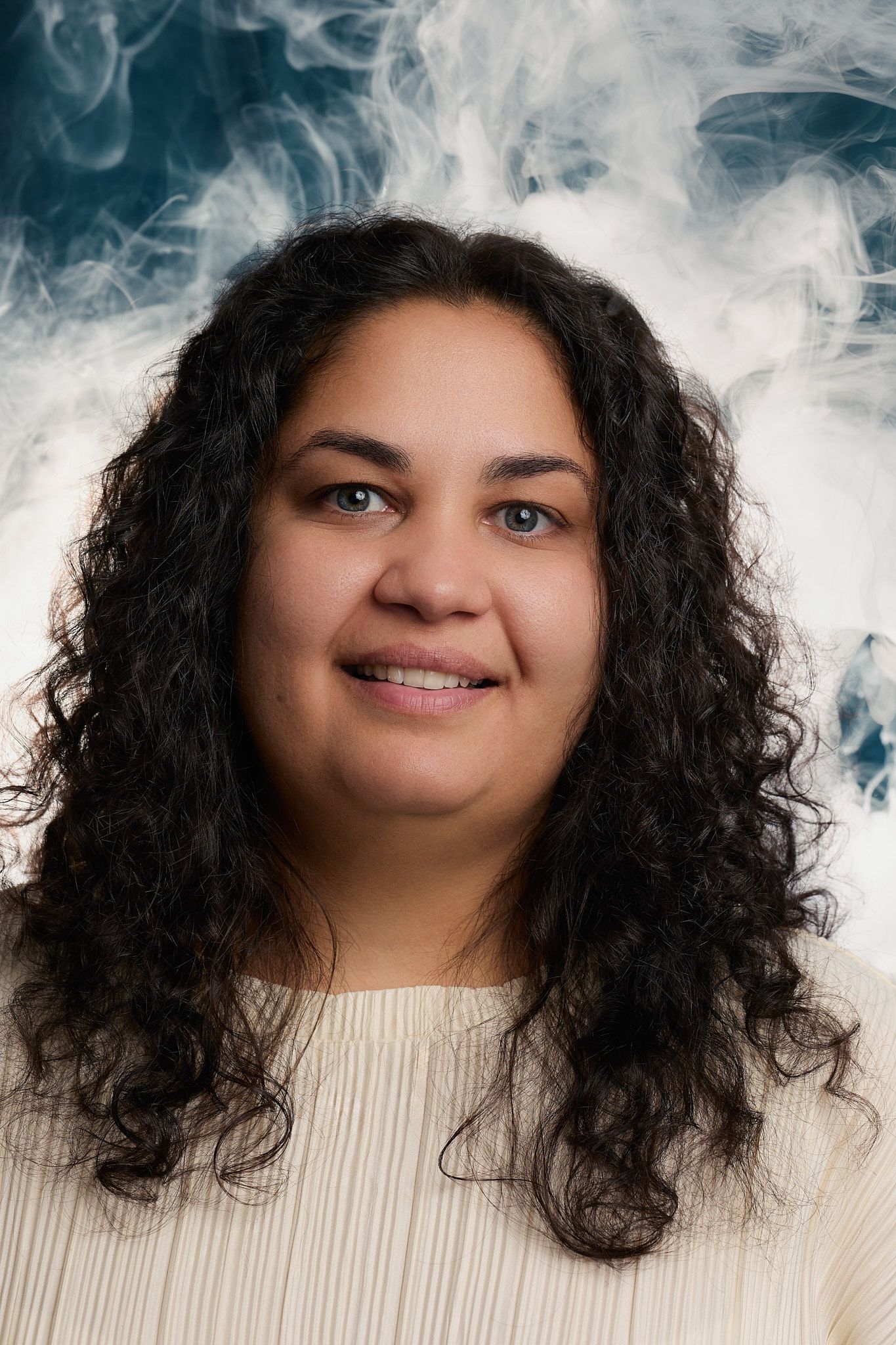 A person with curly dark hair wearing a beige sweater poses against a smoky white background for multiple expressions.