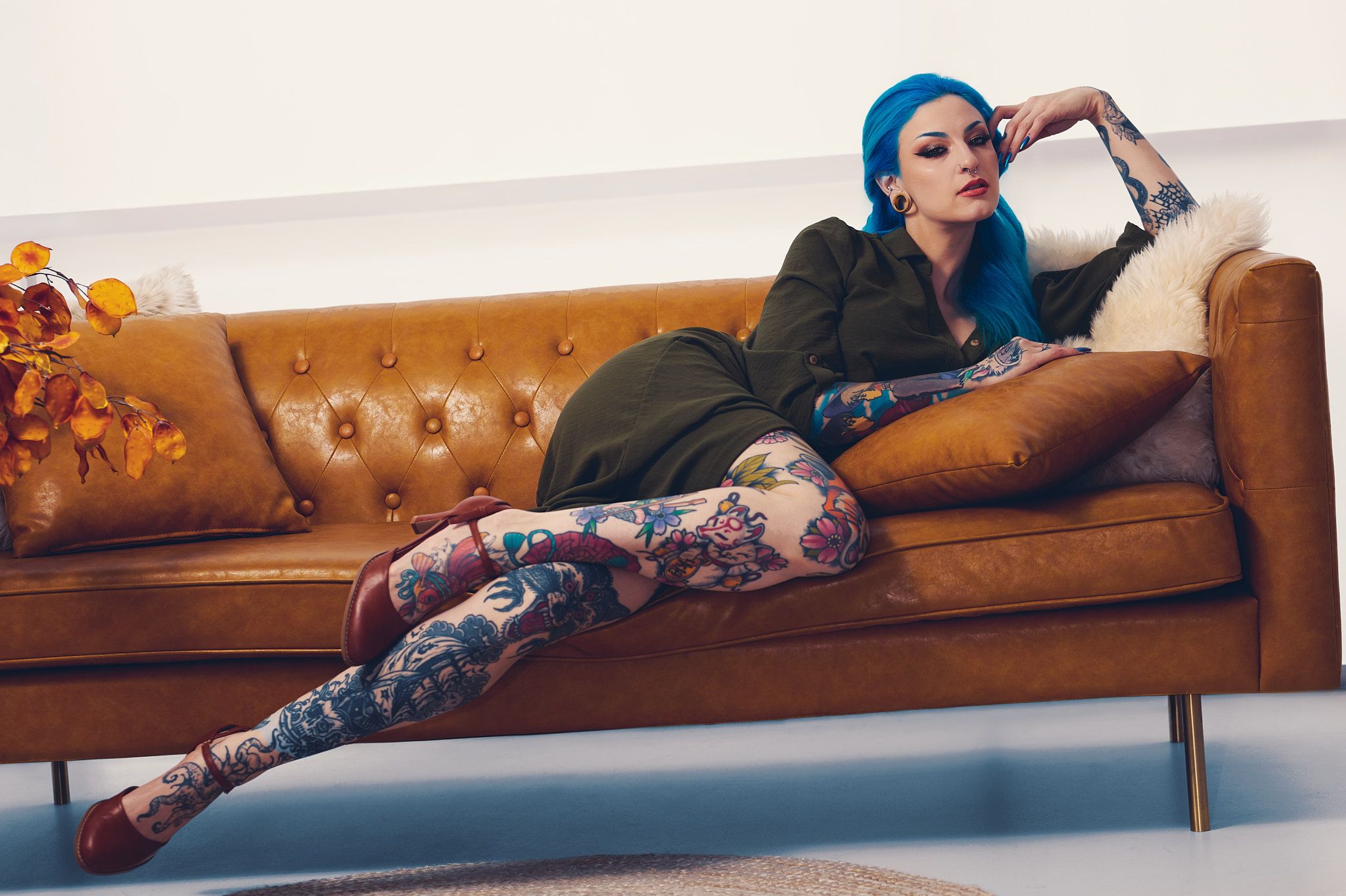 A heavily tattoed woman with blue hair lounges on a brown leather couch