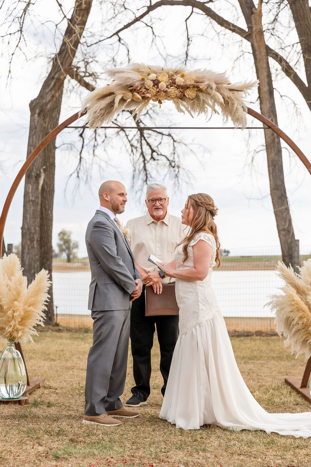 Bride and groom exchanging vows under a circular floral arch with pampas grass outdoors.