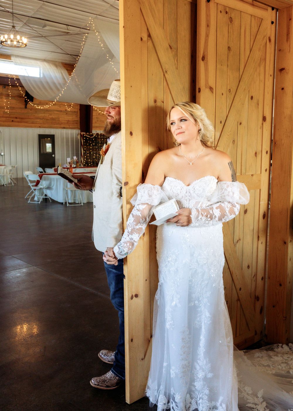 Bride in lace gown holding groom's hand at barn wedding venue door during first look moment.