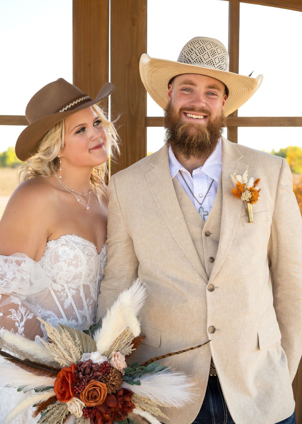 Smiling bride and bearded groom in cowboy hats posing together at rustic country wedding celebration.
