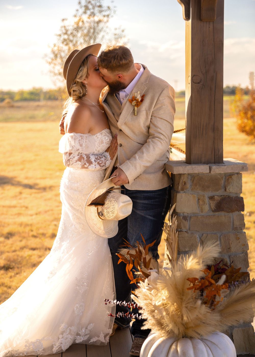 Bride and groom sharing a kiss at golden hour beside rustic stone pillar at outdoor wedding venue.