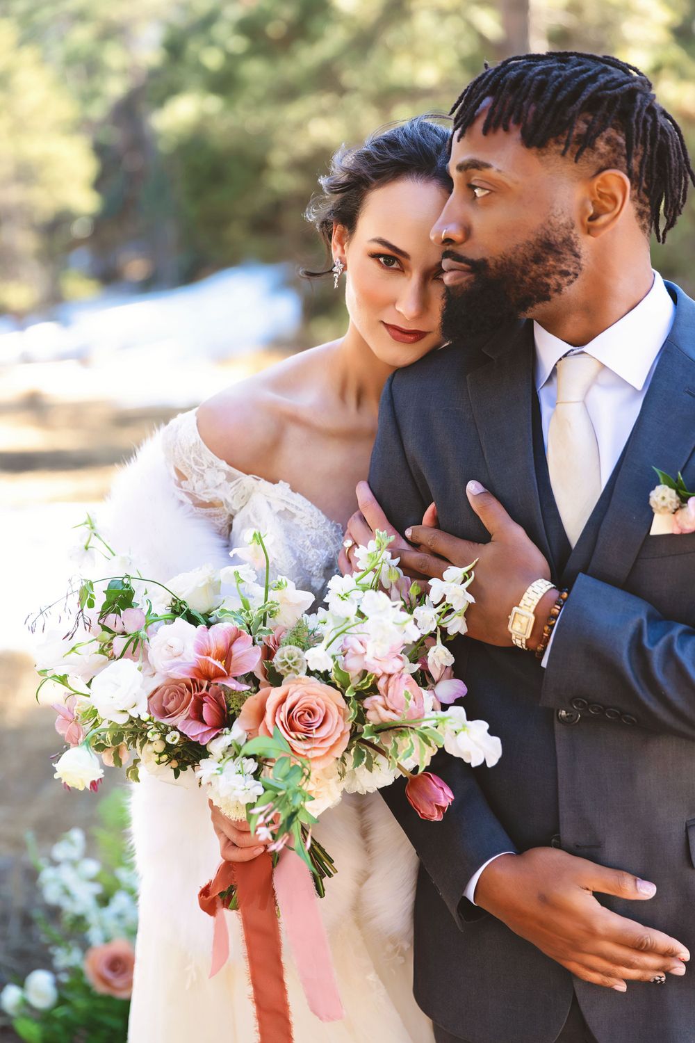 Elegant interracial couple in wedding attire, bride holding pink floral bouquet, groom in gray suit embracing her outdoors.
