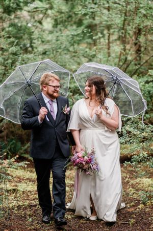 Newlyweds share playful moments under clear umbrellas in a lush forest setting during their rainy Washington elopement