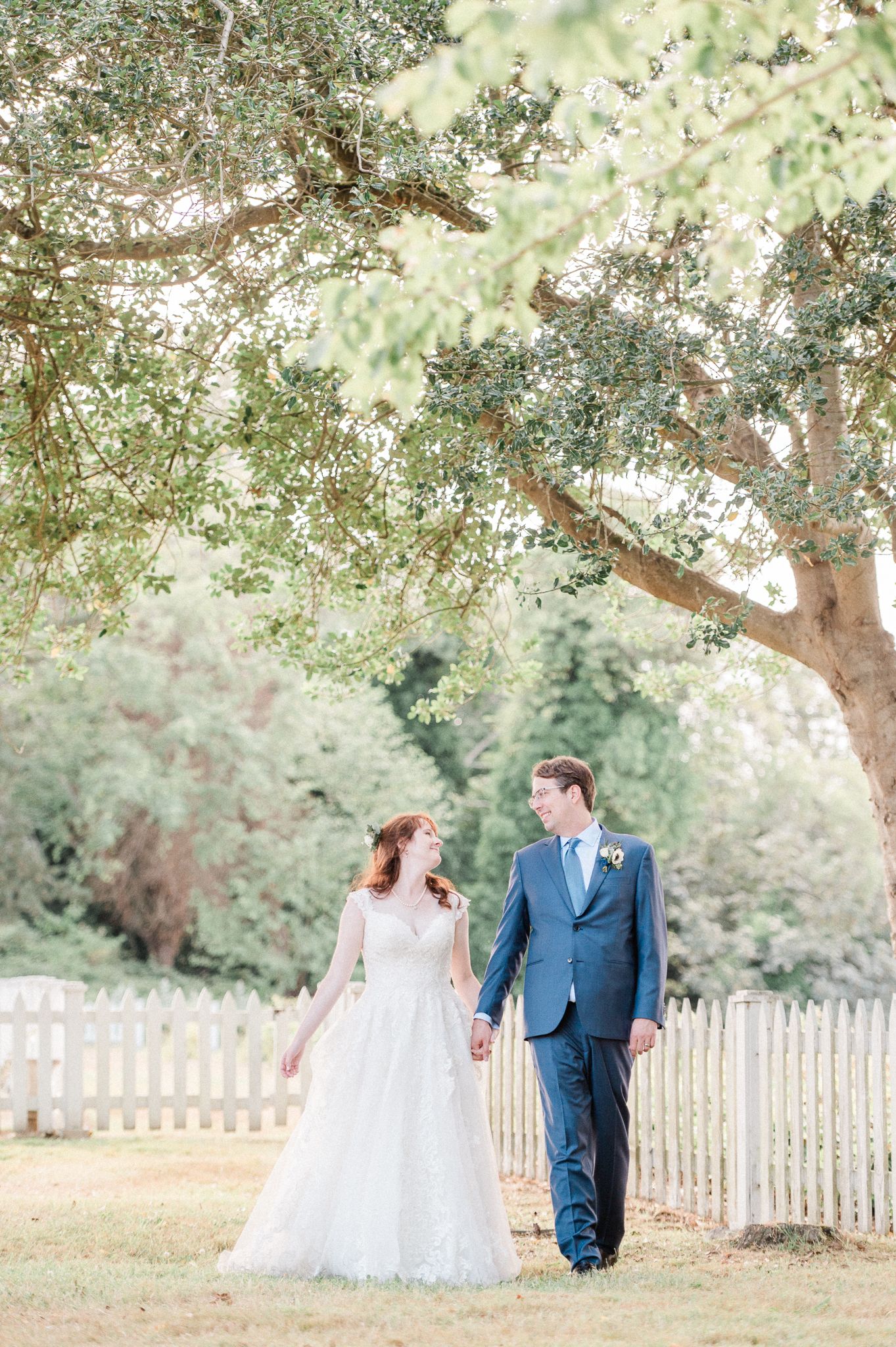 A couple in wedding attire walks together under sunlit trees in a garden setting with a white picket fence during their Port Gamble wedding