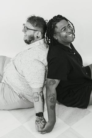 Two tattooed people sitting back-to-back smiling, wearing glasses, in a black and white studio photo.
