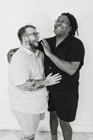 Two friends laughing together in black and white photo, one with tattoos and glasses, the other with dreadlocks and glasses.