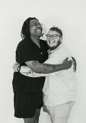 Two friends laughing and hugging joyfully in a black and white portrait photo, both wearing glasses and casual outfits.