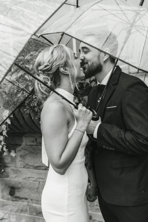 A romantic black and white wedding photo of a couple sharing an intimate moment under a clear umbrella.