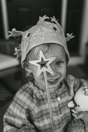 Child wearing a crown and holding a star wand smiles in black and white portrait.