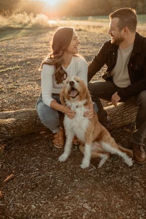Couple sits with their brown and white dog on a log during golden hour sunset.