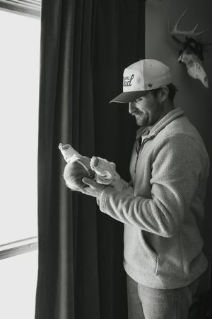 A person in a baseball cap and sweatshirt stands by a window holding two small birds in their hands in black and white.