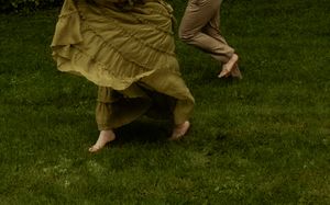 Two barefoot dancers in earthy tones perform on lush green grass, skirts swirling in graceful motion.