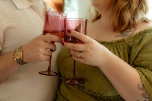 Close-up of couple clinking pink stemmed wine glasses, woman wearing engagement ring and green dress.