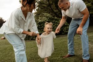 Family helps young child take steps on grass in casual outdoor setting.