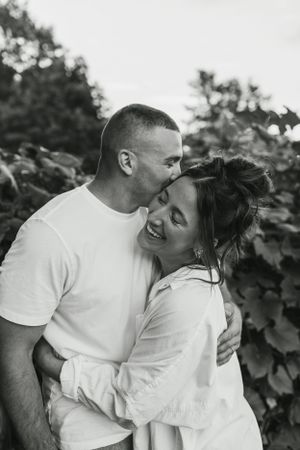 A tender black and white portrait of a couple embracing outdoors against foliage background.