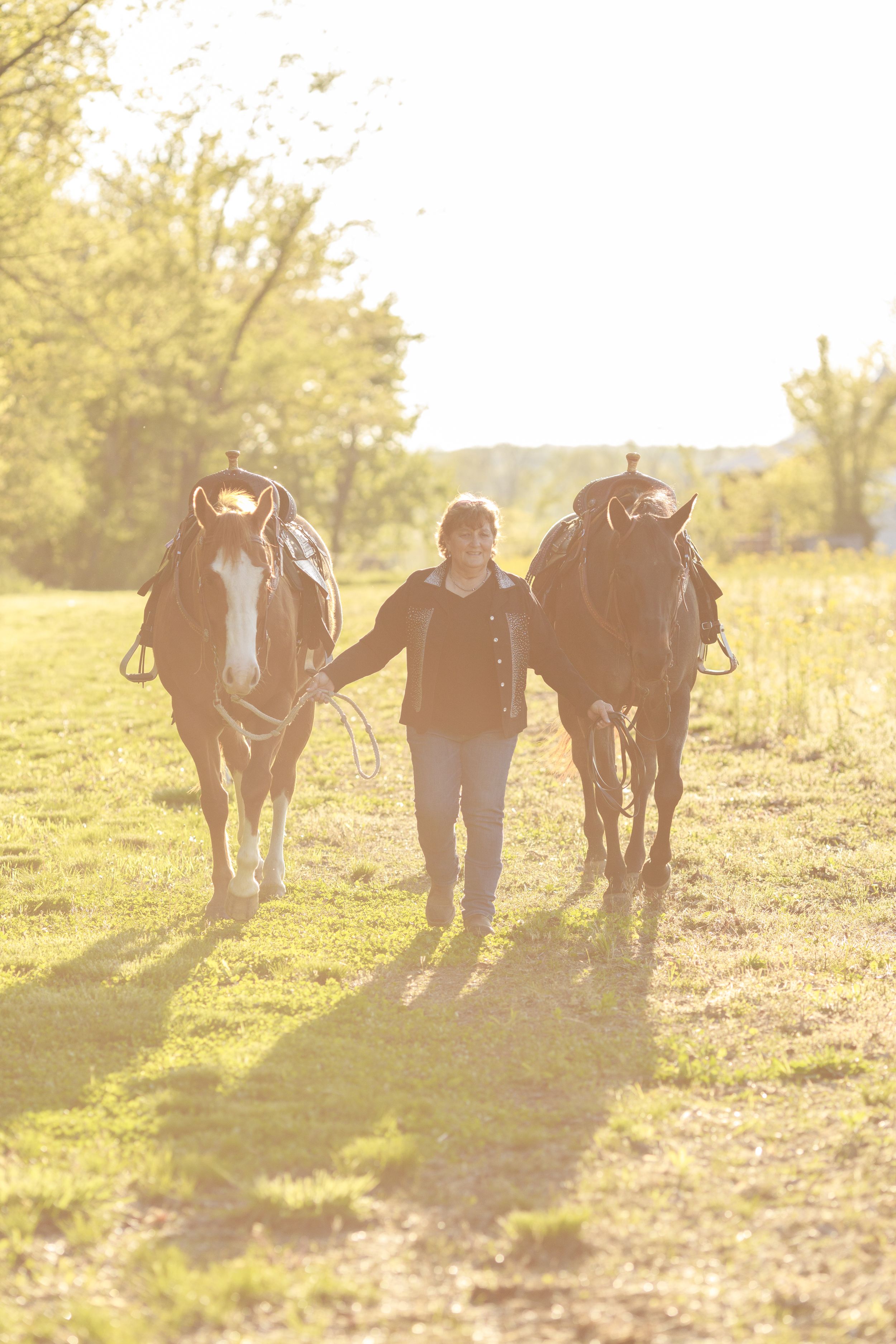 A cowgirl walks her two horses in a blast of evening light.