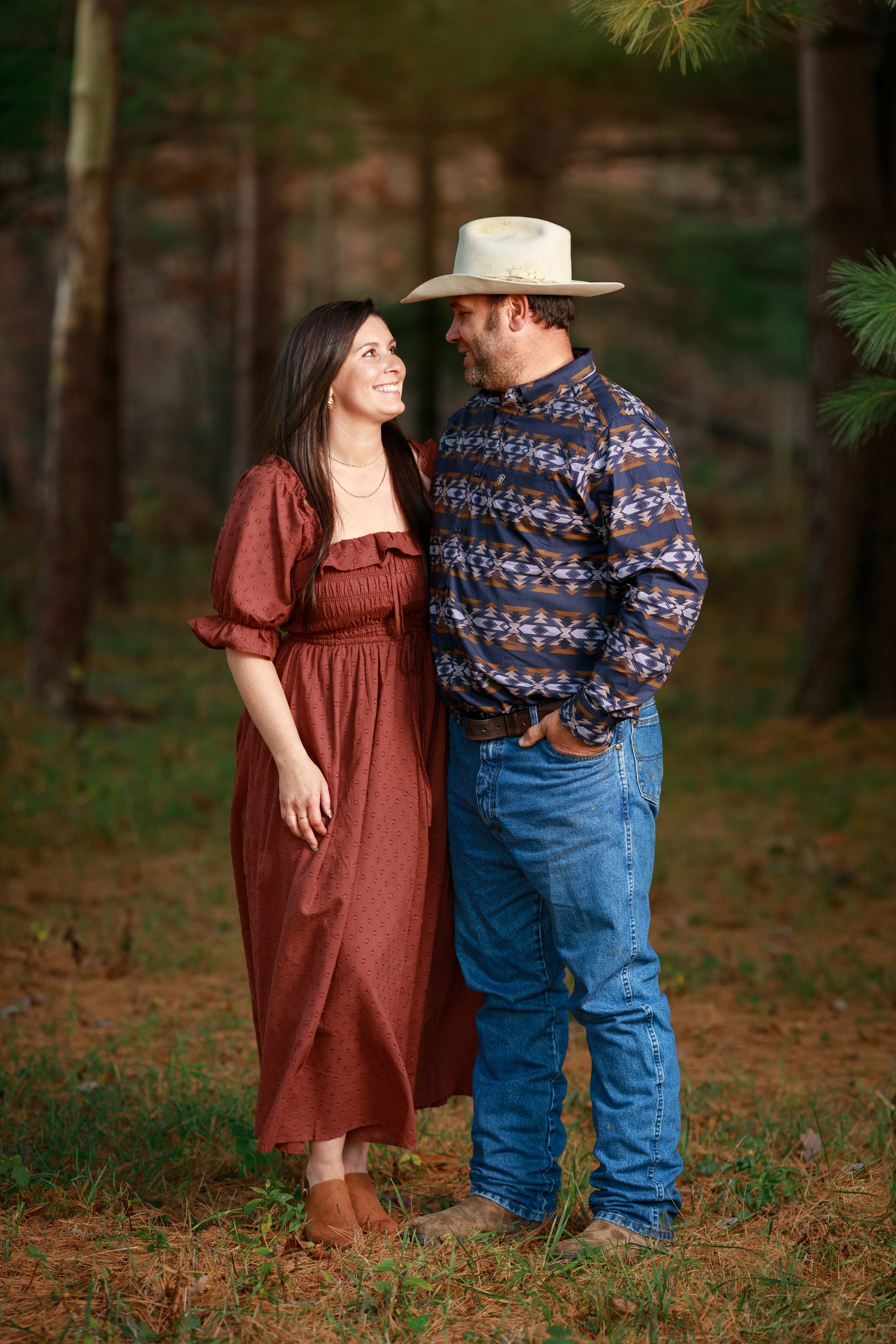 Married couple poses in the Helm's Tree Farm pines. They are wearing fall colors and western attire and are staring lovingly at each other.