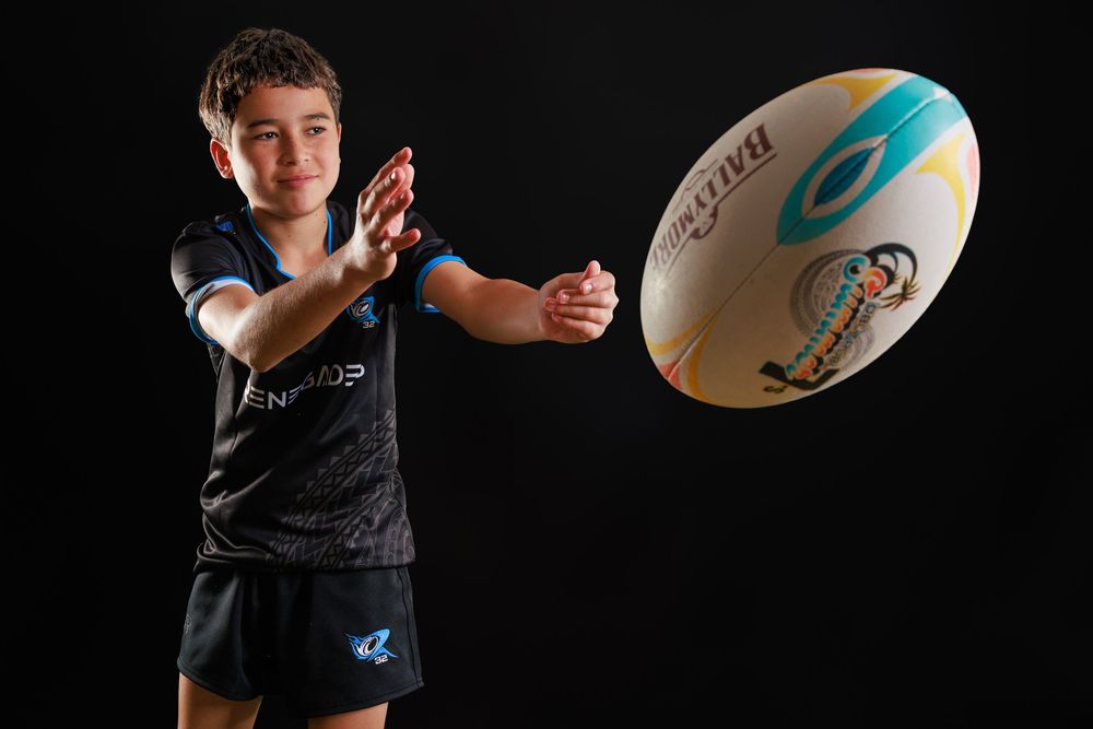 Young boy in a black and blue rugby kit smiling as he passes a Gilbert rugby ball against a dark studio background.