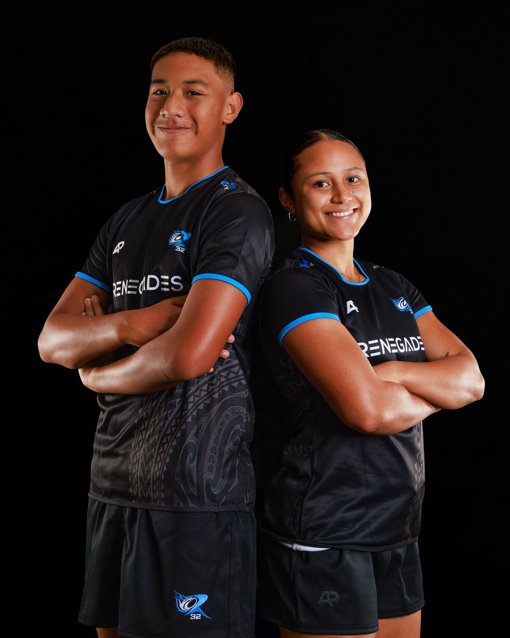 Two smiling teammates stand back to back with arms crossed, wearing black and blue Renegades sports jerseys and shorts against a black backg