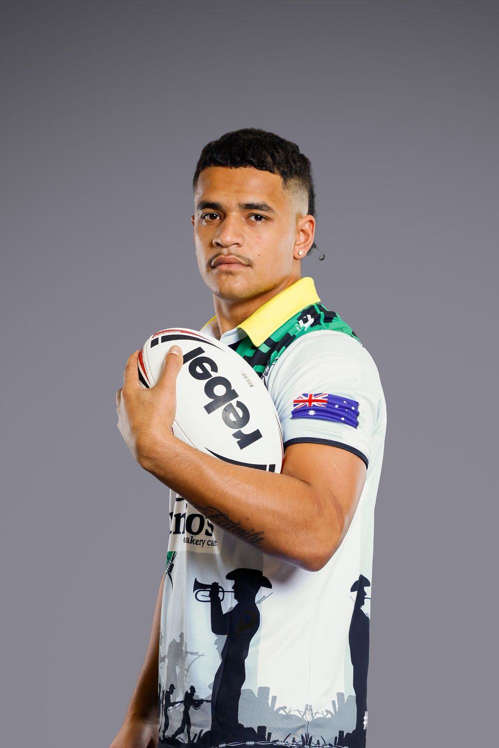Rugby league player holds a Rebel football in a white commemorative jersey with a bugler silhouette, yellow collar and Australian flag.