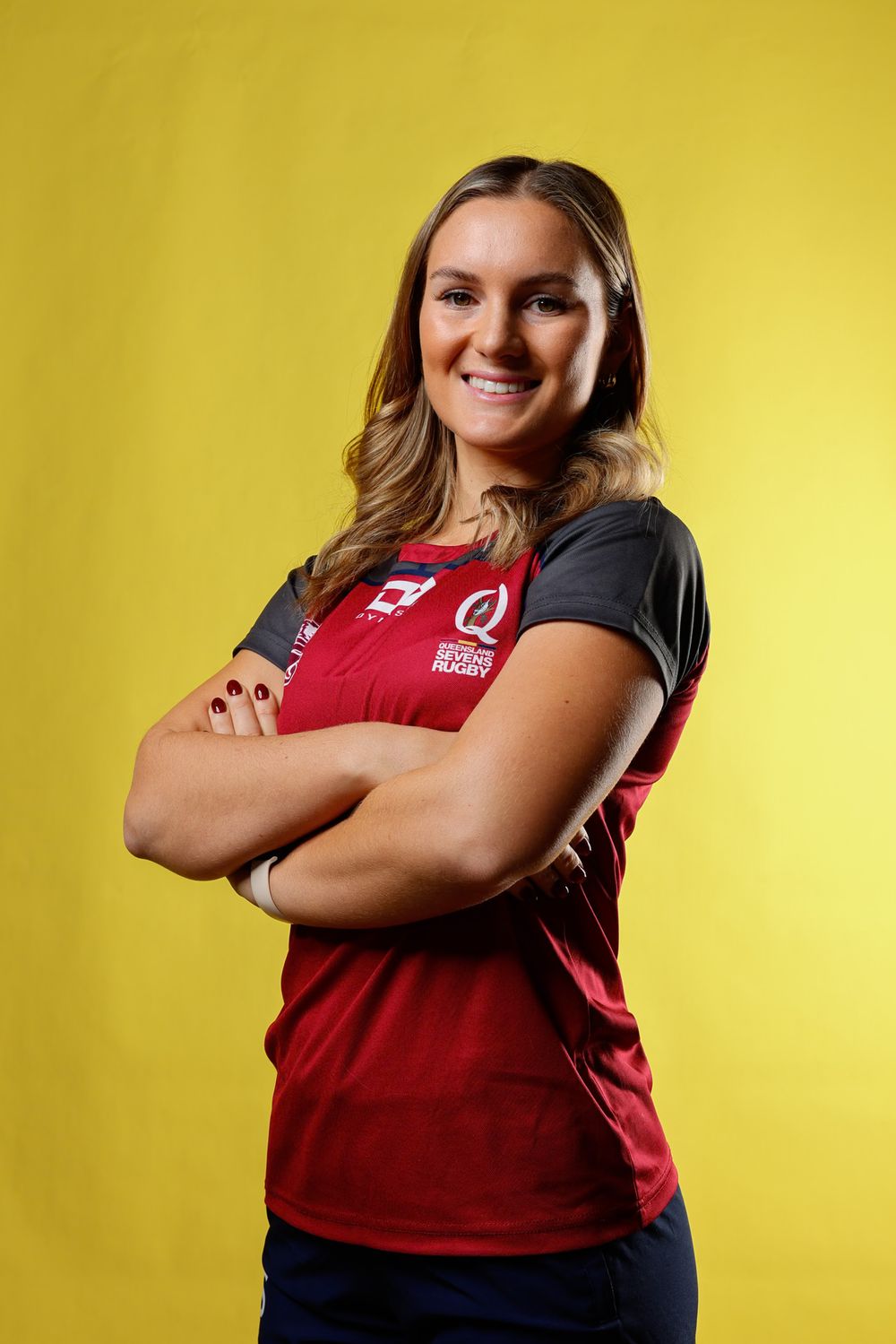 Smiling woman with wavy hair and arms crossed wears a maroon and gray rugby jersey while posing against a bright yellow studio background.