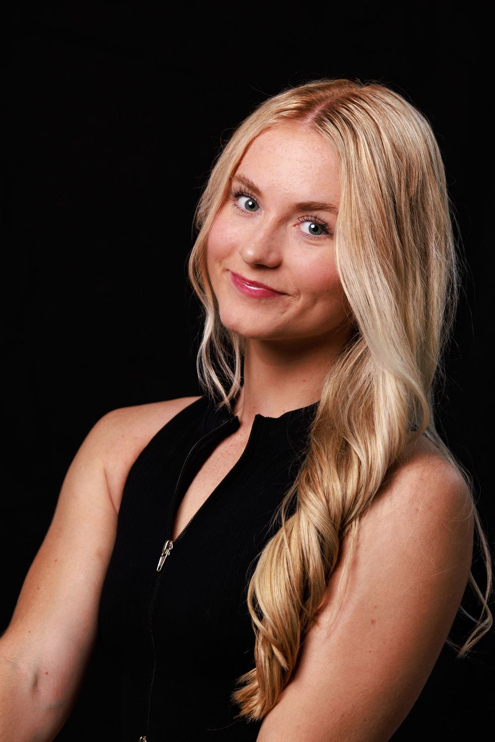Smiling woman with long wavy blonde hair wearing a black zip-up top against a dark studio background.