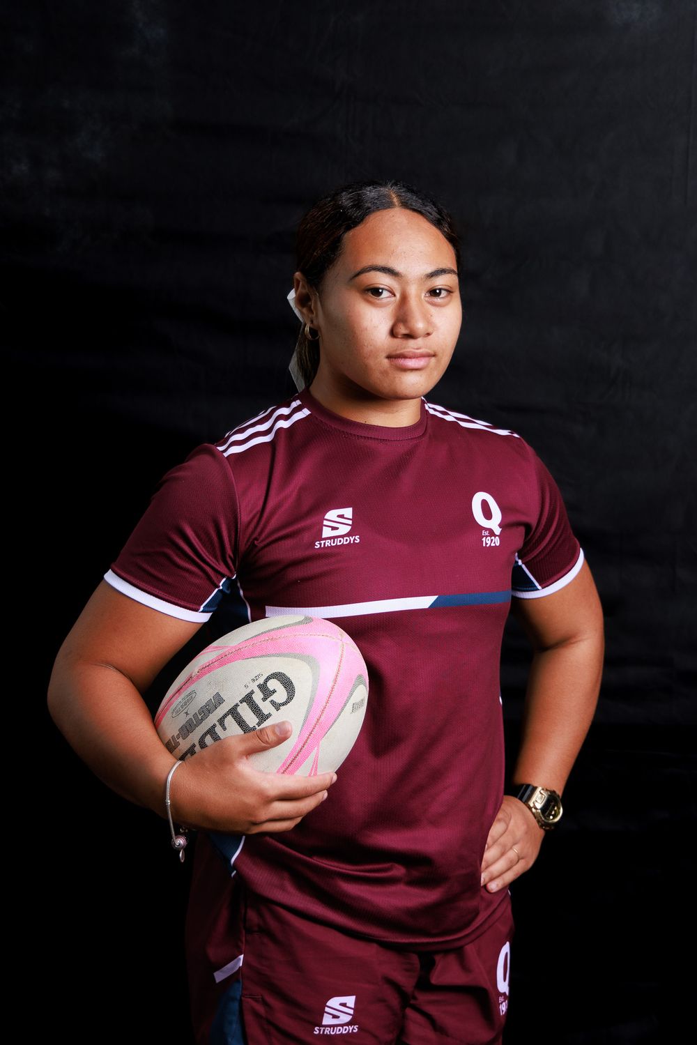 Rugby player in a maroon Queensland jersey holding a pink and white Gilbert rugby ball against a dark background.