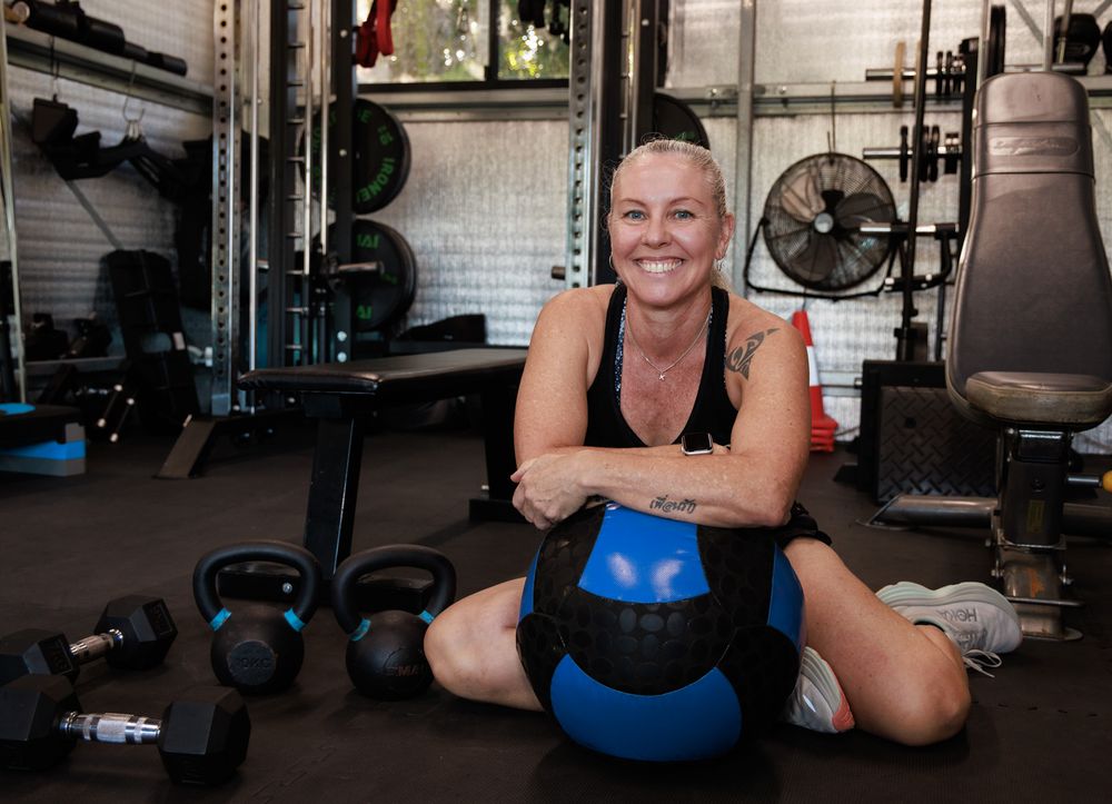 Smiling woman with short blonde hair sits on a gym floor hugging a blue medicine ball beside kettlebells and dumbbells.