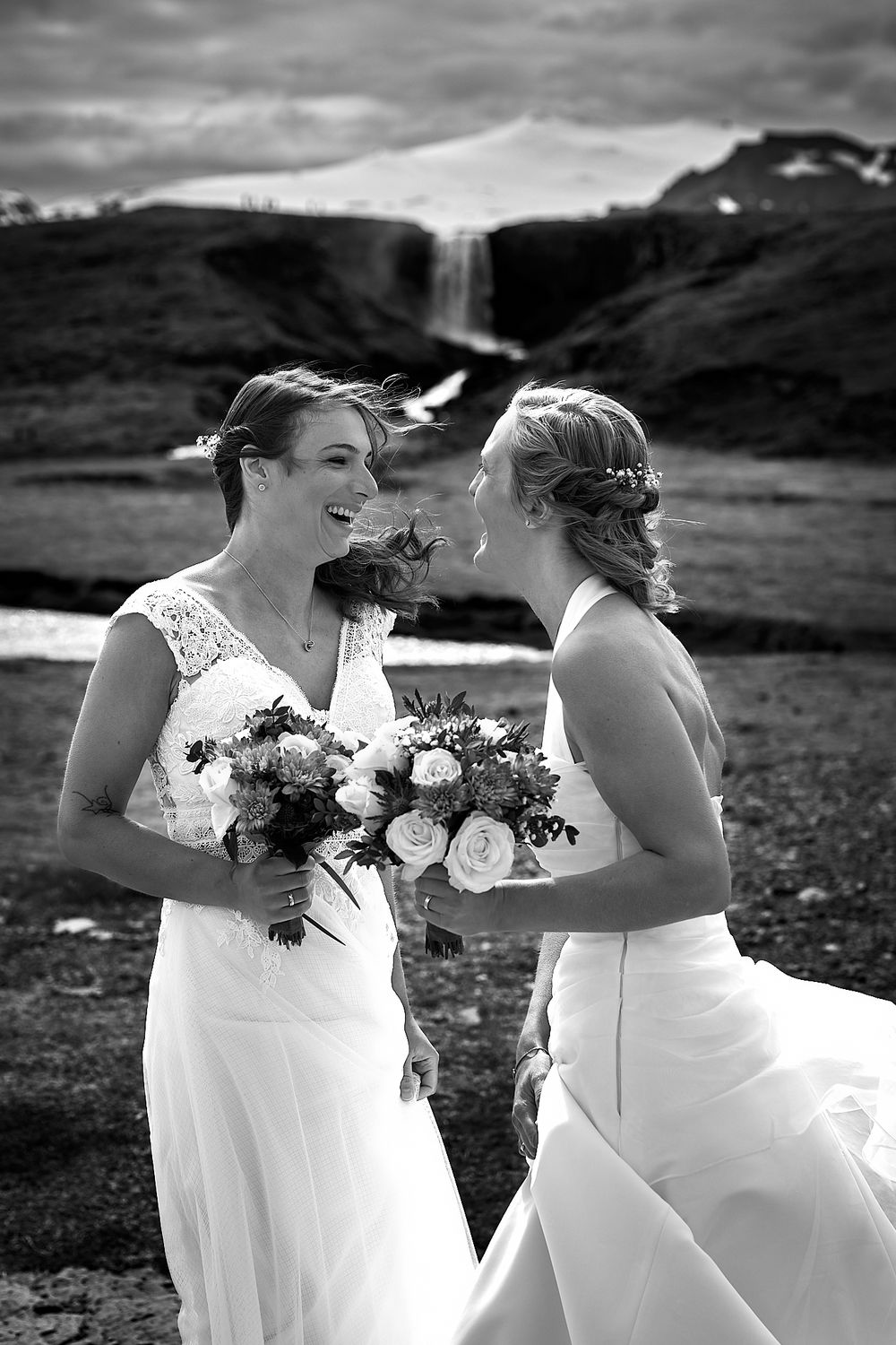 A black and white portrait of two brides sharing a joyful moment with bouquets against a mountain backdrop.