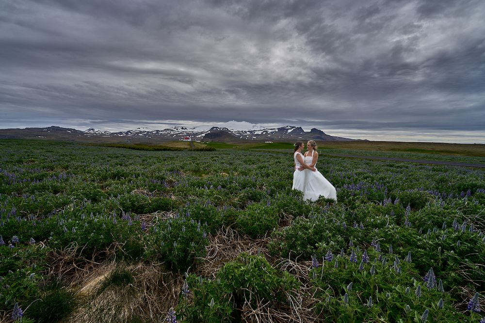 Two brides embrace in a dramatic landscape with stormy skies and mountains.