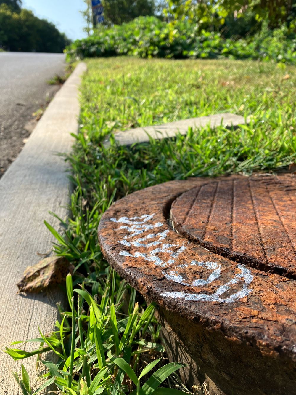 Rusty manhole cover with graffiti writing surrounded by grass and concrete.