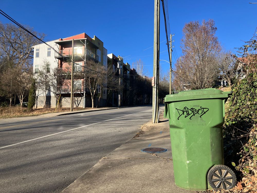 A green garbage bin sits on a suburban street curb near a bush on a sunny day.