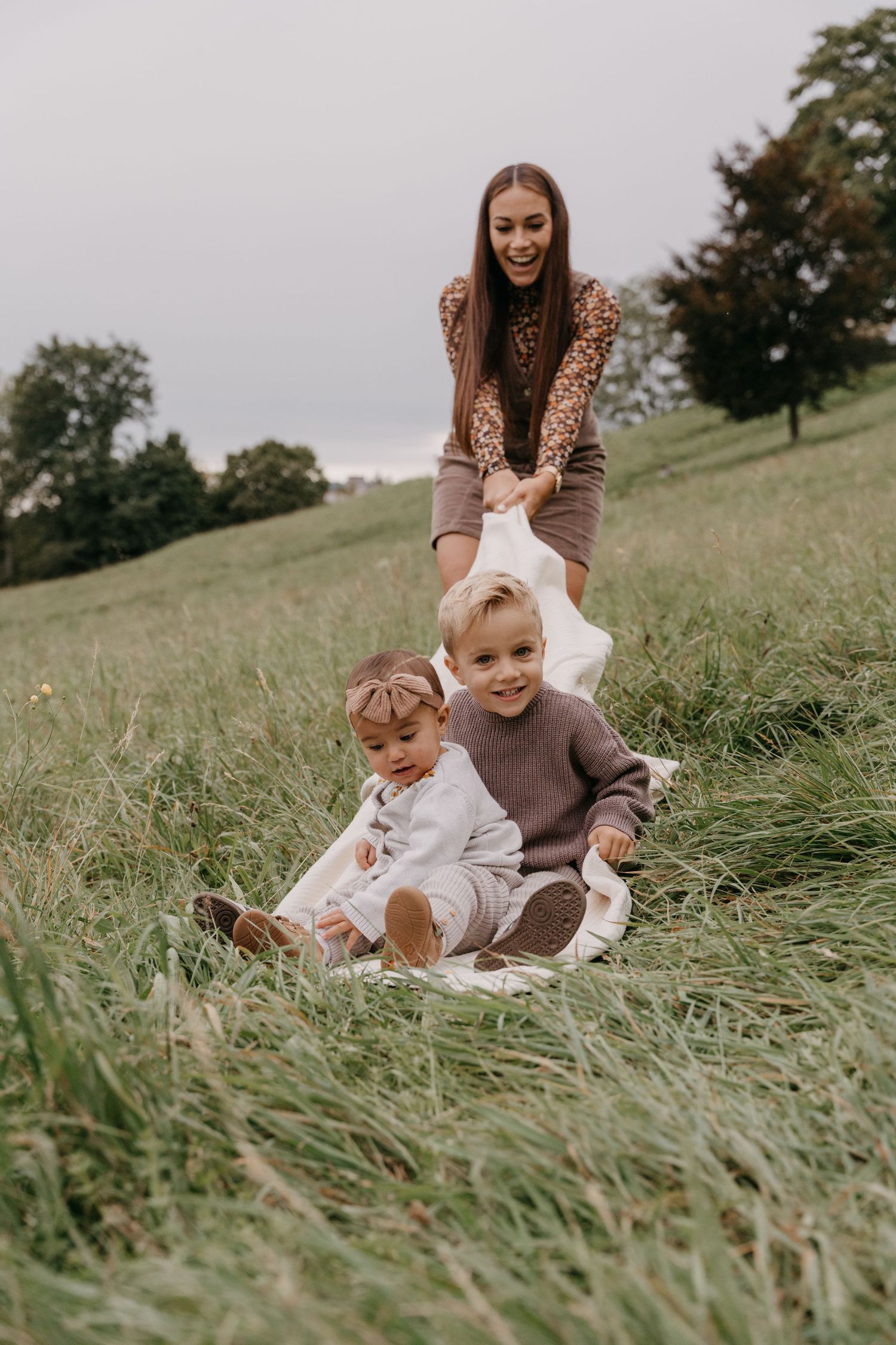 Family sitting on white blanket in grass field enjoying playful candid moments together.