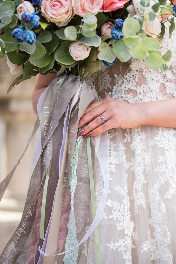 Close-up of bridal bouquet with pink roses, blue flowers and flowing grey ribbons against lace dress.