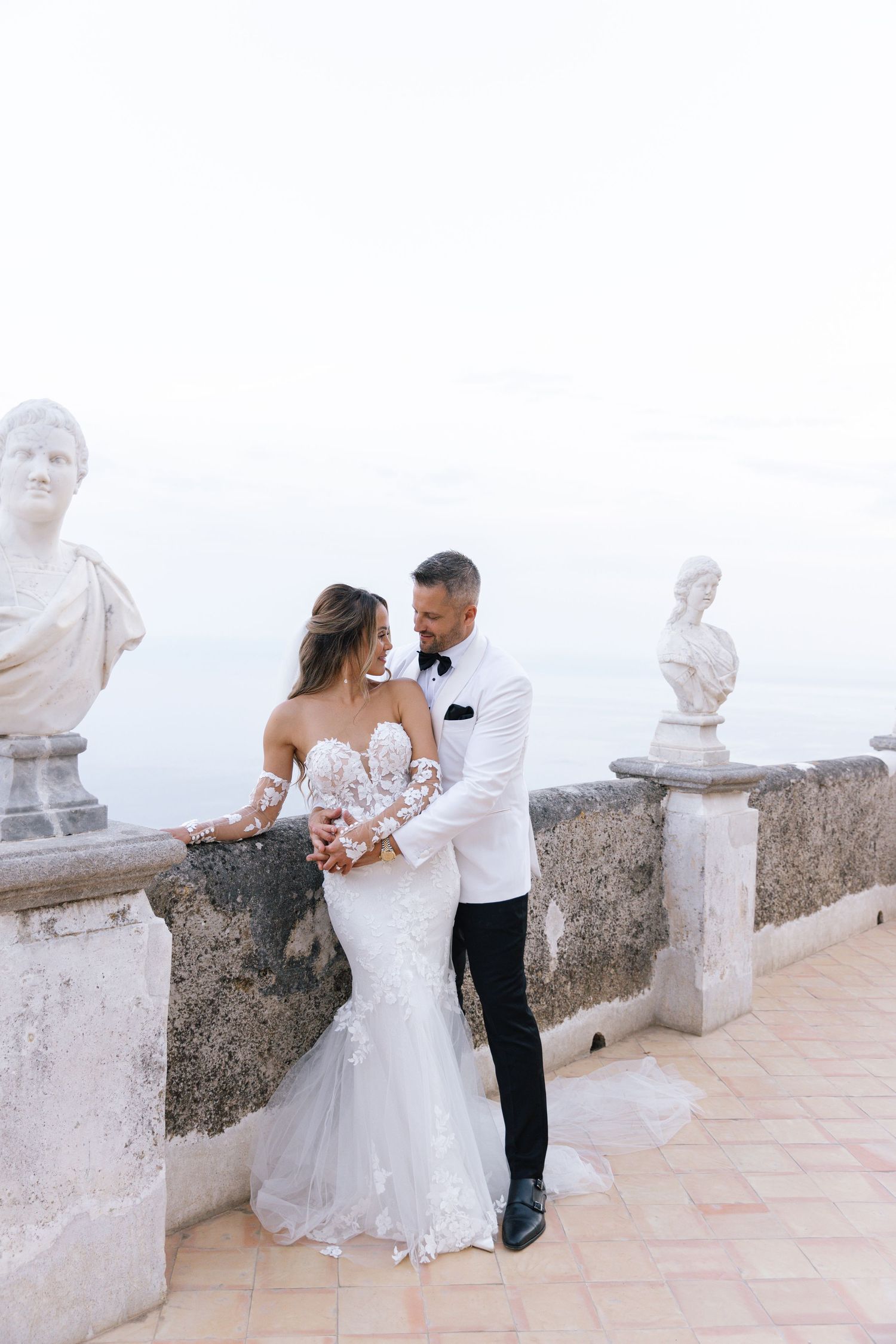 Bride in lace gown poses with white bouquet next to classical stone statue against ocean backdrop.