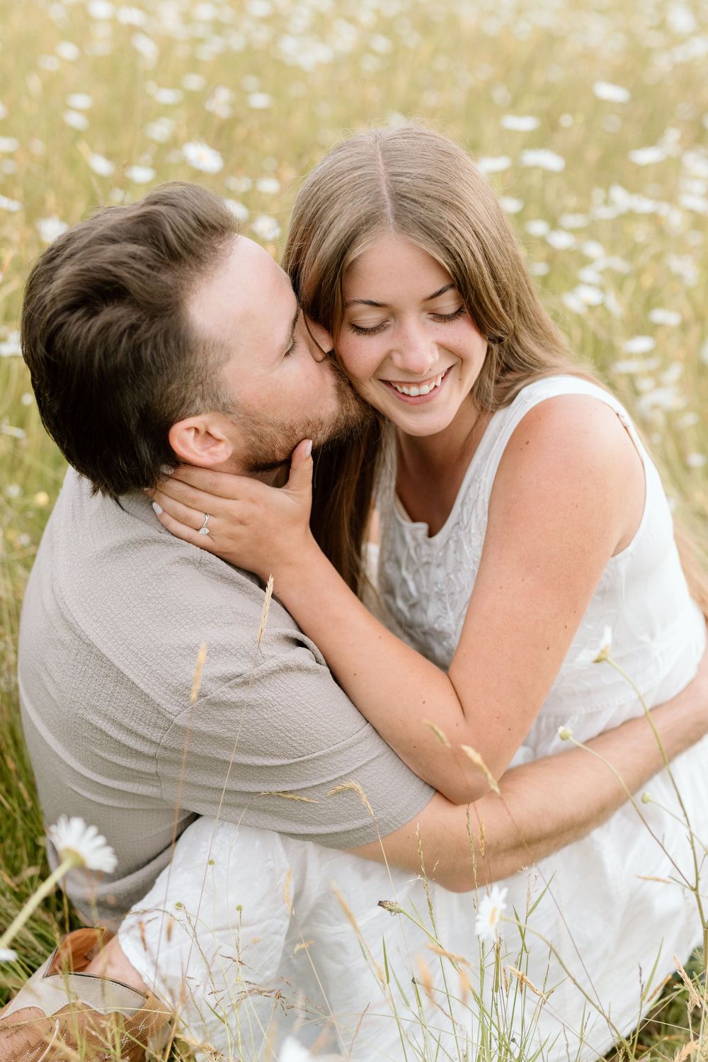 A couple shares tender moments in a sunlit meadow of daisies, reclining together in white summer outfits.