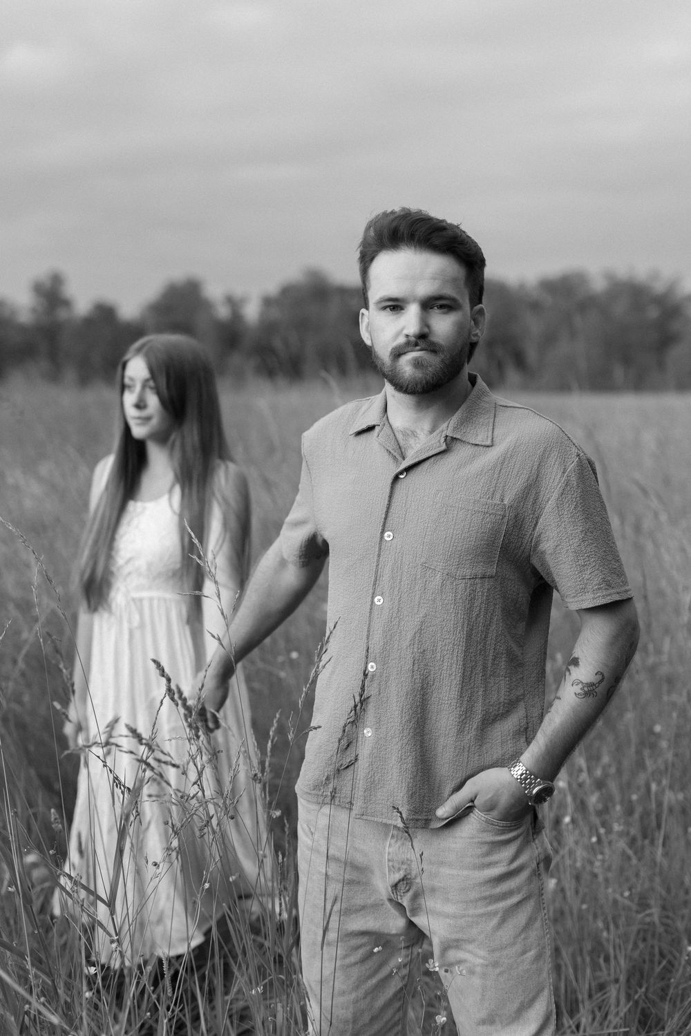 A couple stands together in a field of tall grass and wildflowers in a black and white artistic portrait series.