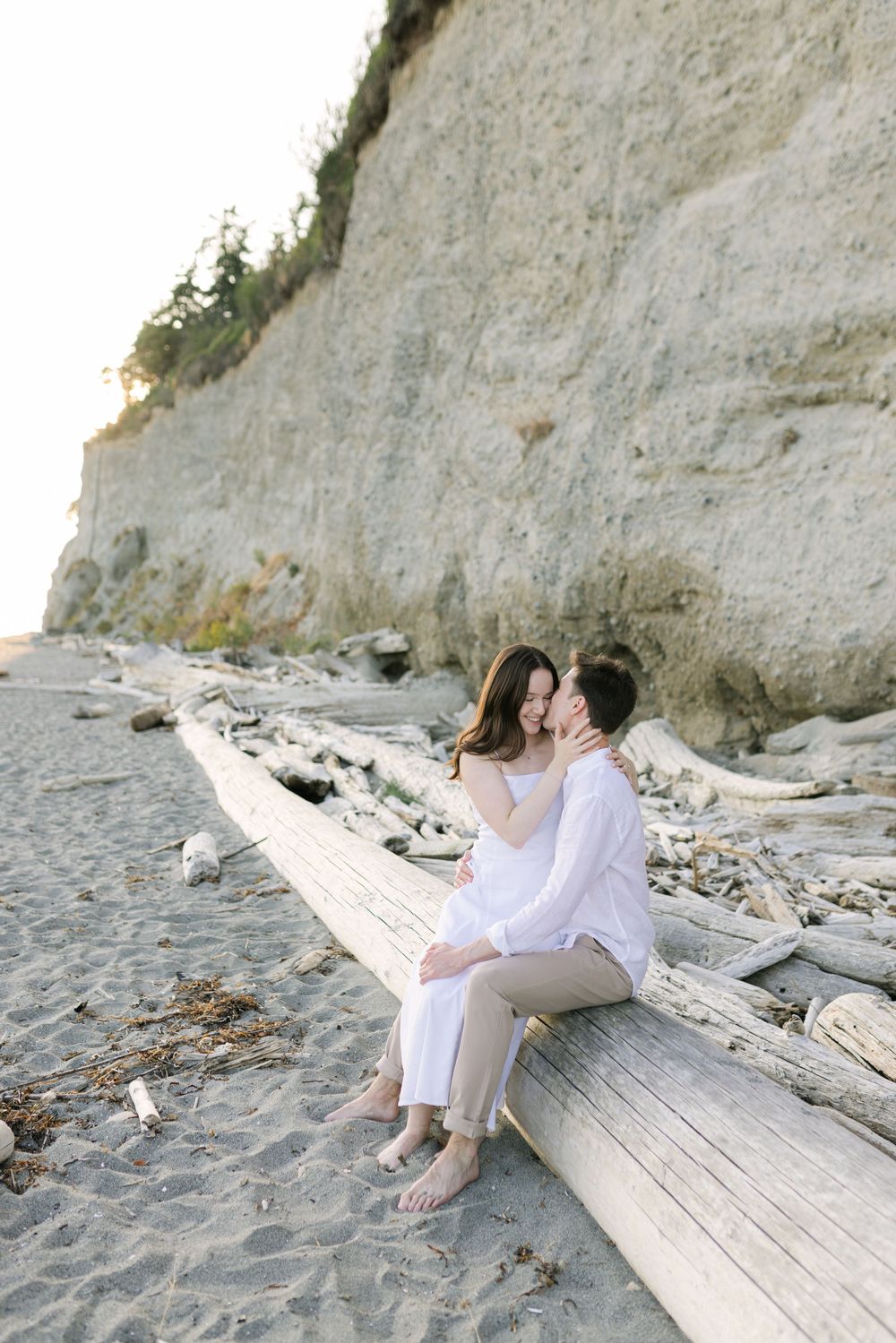 Couple sits together on driftwood log at beach with dramatic white cliffs in background during sunset.