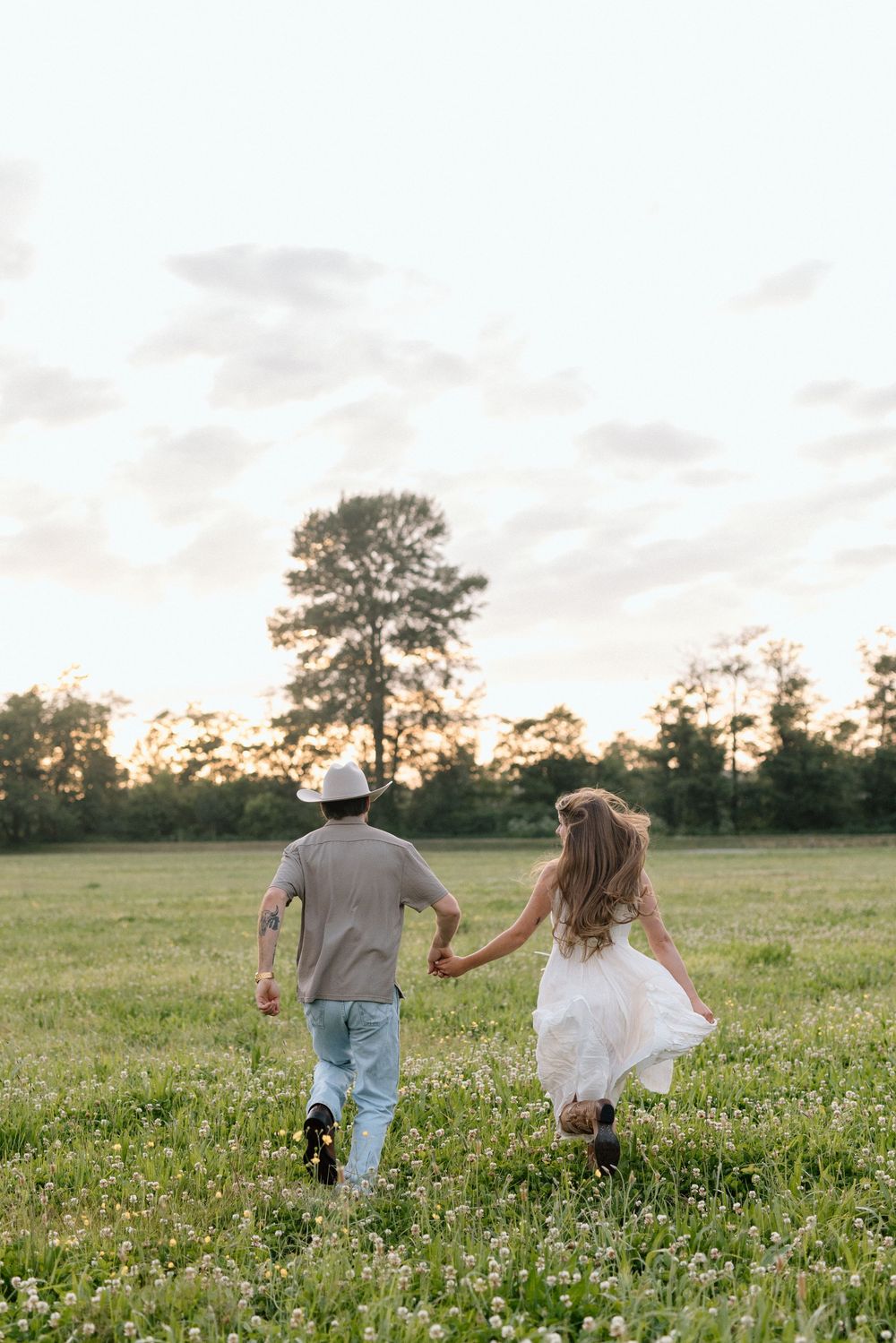 A couple runs hand in hand through a wildflower meadow at sunset with tall trees in the background.